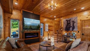 Interior acolhedor de uma cabana de madeira com lareira, sofás de couro, uma televisão e uma escada.