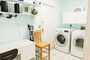 A laundry room with two washing machines, a chair, a desk, and shelves with various items. -  Three Palms 3BR / sleeps 8 / beach 5 min. / backs to nature trail & park w/lake (Largo)