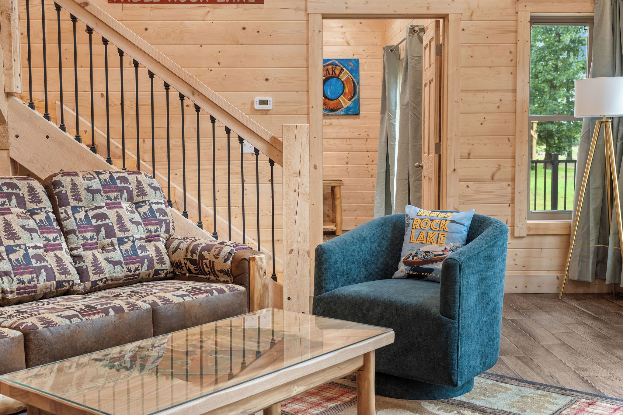A wooden interior with a sofa, a blue armchair, a glass coffee table, and a staircase.