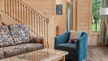 A wooden interior with a sofa, a blue armchair, a glass coffee table, and a staircase.