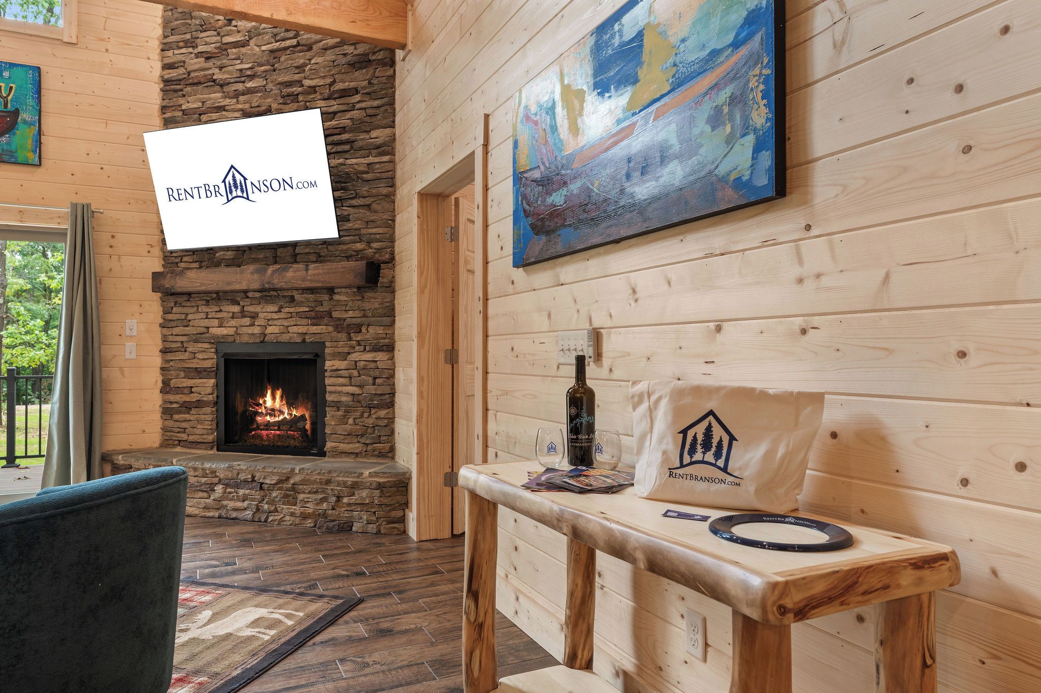 A cozy interior with a stone fireplace, wooden walls, a table with a bottle and glasses, and a painting on the wall.