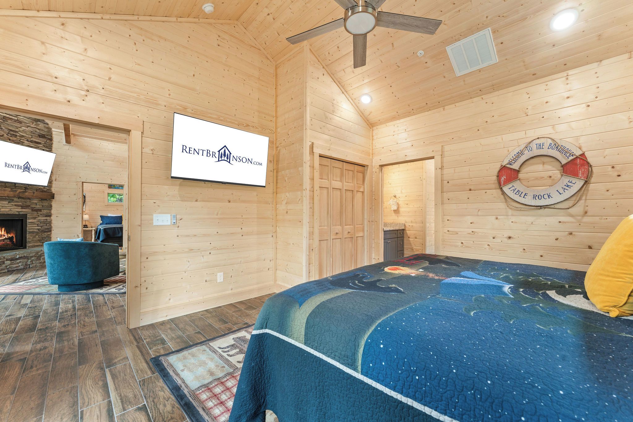A wooden cabin interior with a bed, a fireplace, a ceiling fan, and a sign for RentBrinson.com.