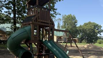 A wooden play structure with a slide and climbing area, surrounded by trees and gravel.