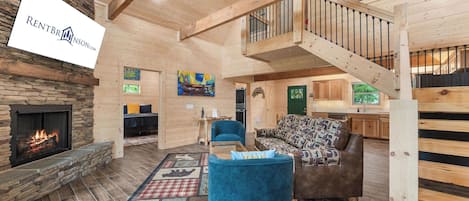 A spacious living area with a stone fireplace, a blue sofa, a patterned rug, and a wooden staircase.