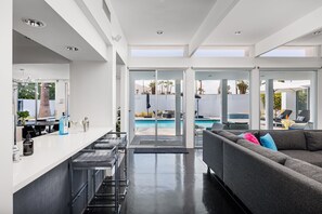 A modern kitchen with a bar area, a pool, and a living room with a sofa. - The Bond House | 37-Ft Salt Pool, Mosaic Spa, Fire Pit, Outdoor Shower, BBQ (Palm Springs)