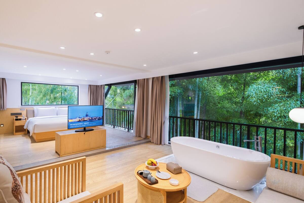 A modern hotel room with a large bed, a TV, a bathtub, a balcony with a view of greenery, and a dining area with a table and chairs.