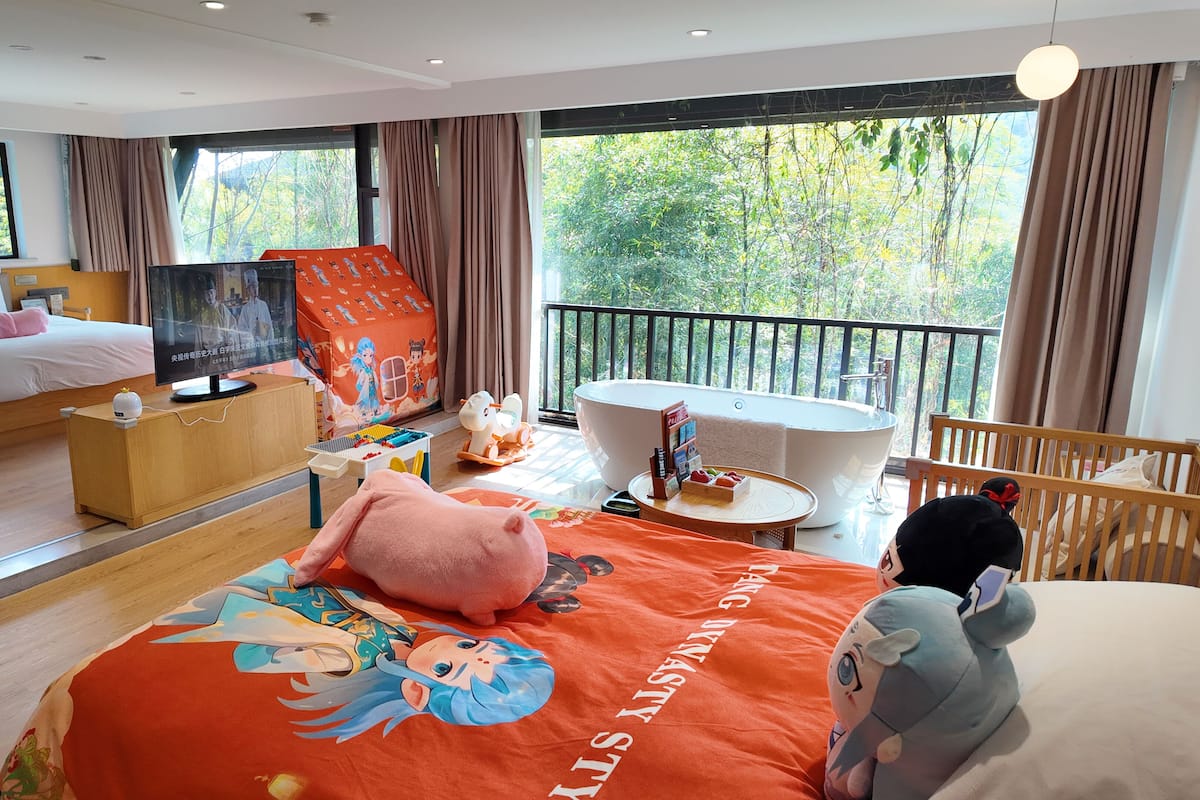 A hotel room with a large bed, a TV, a bathtub, and a balcony with a view of trees.