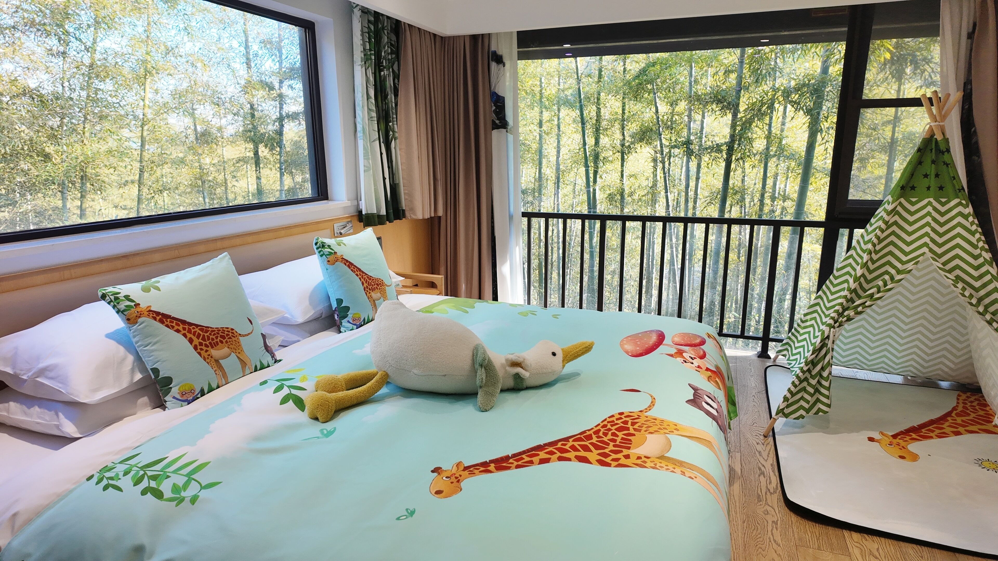 Habitación de hotel con una cama y ropa de cama con temática de jirafa, un ventanal con vistas a árboles y suelo de madera.