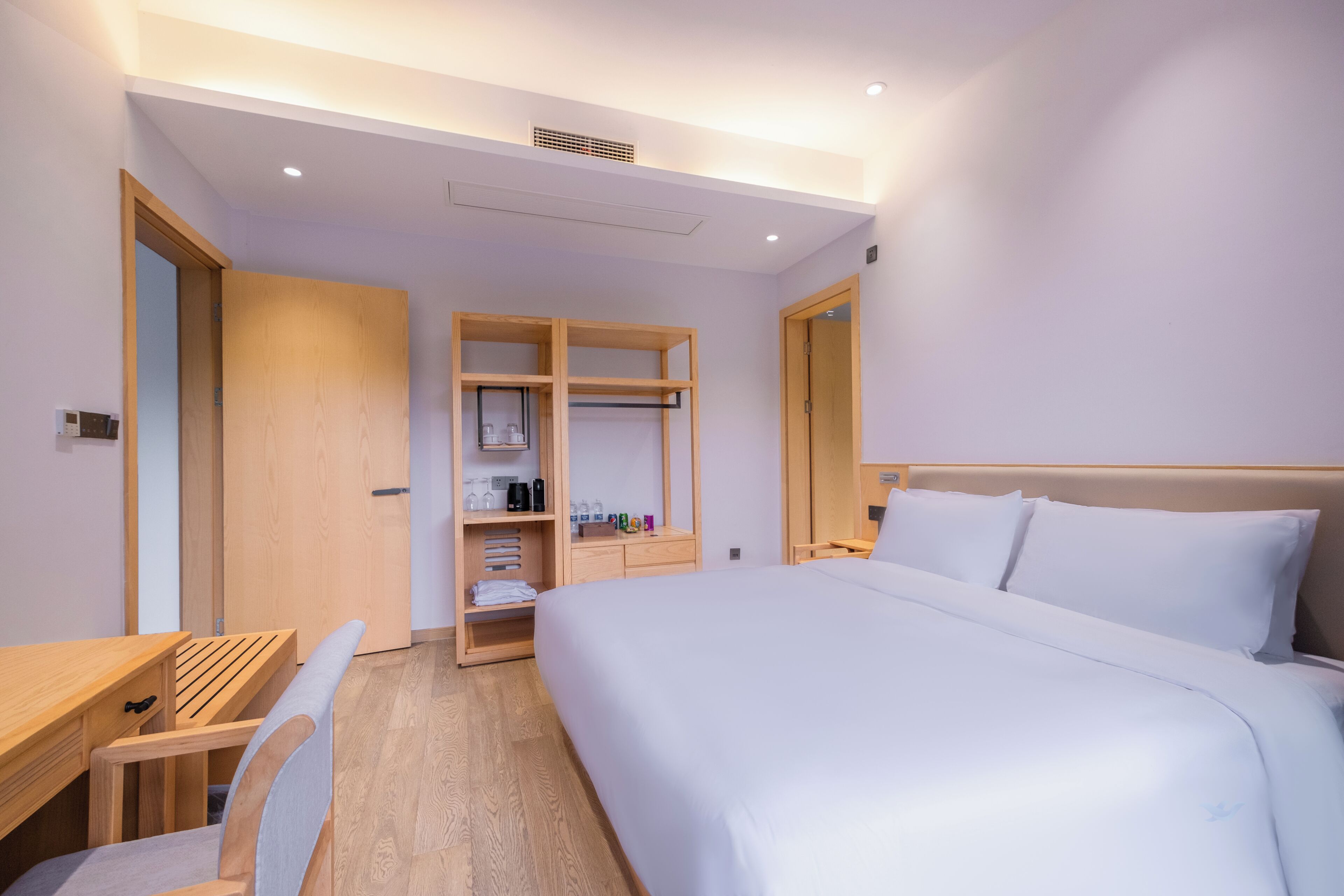 A hotel room with a bed, a desk, a chair, a wardrobe, and a shelf with toiletries.