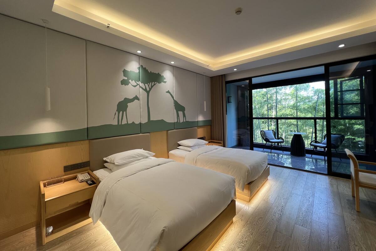 A hotel room with two beds, a desk, a chair, and a large window with a view of trees.