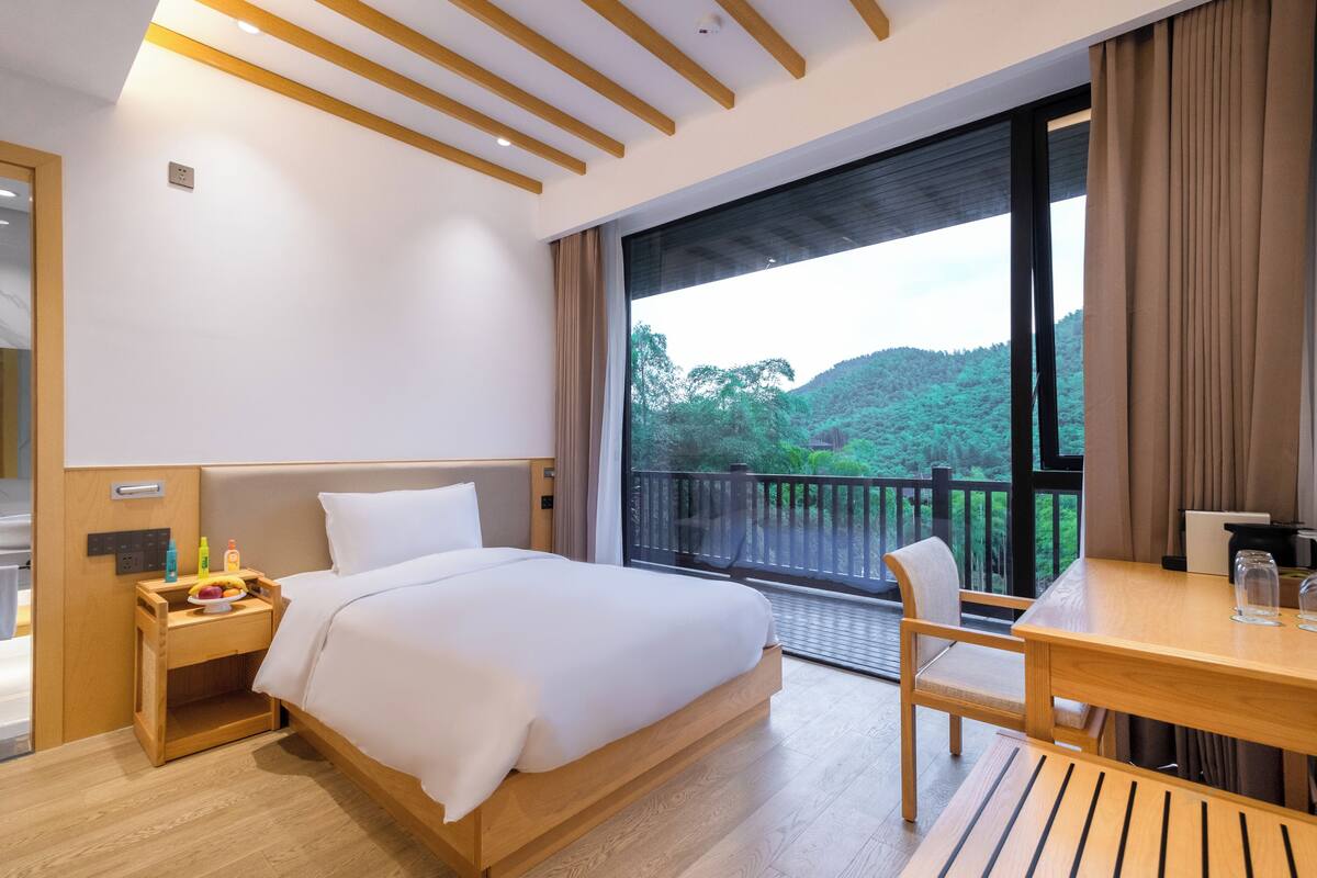 A modern hotel room with a large bed, a wooden nightstand, a dining table, and a balcony with a view of greenery.
