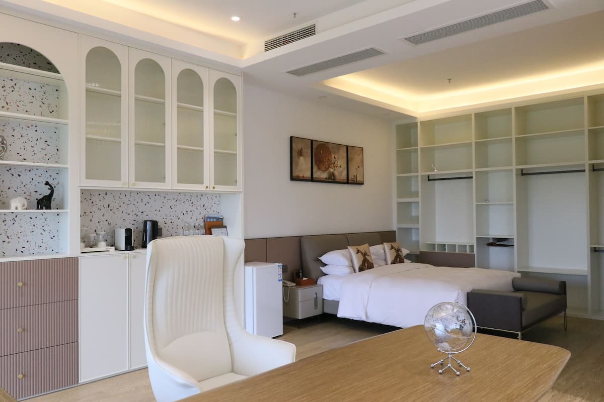 A modern hotel room with a bed, a desk, a chair, a small refrigerator, and built-in shelving.