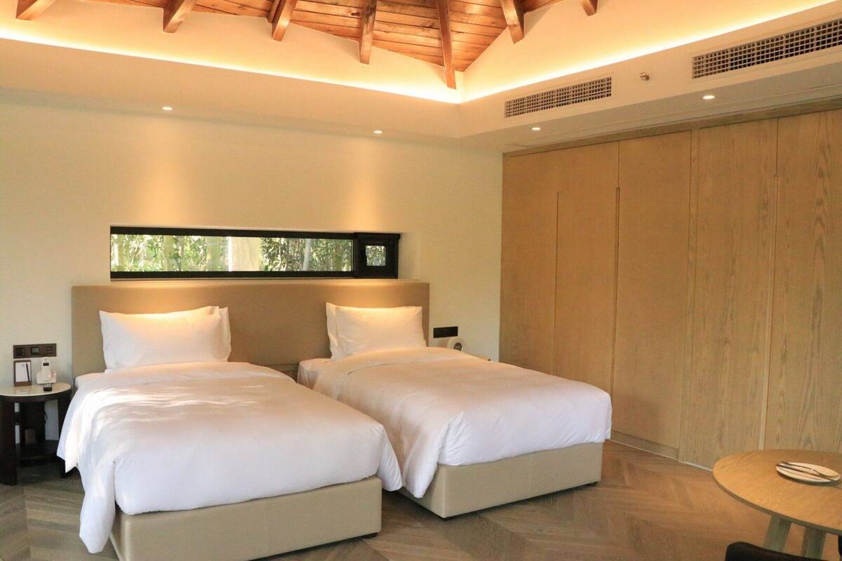 A modern hotel room with two beds, a wooden ceiling, a large window, and a wooden wardrobe.