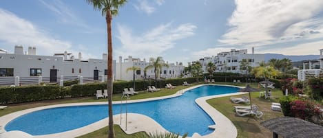 Una piscina de diseño curvo, rodeada de sillas de descanso y palmeras, con edificios al fondo.