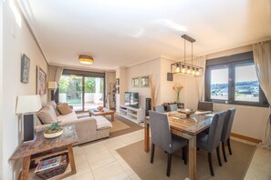 Ein modernes Wohnzimmer mit Sofa, Couchtisch, Fernseher, Esstisch, Stühlen und Blick ins Grüne.