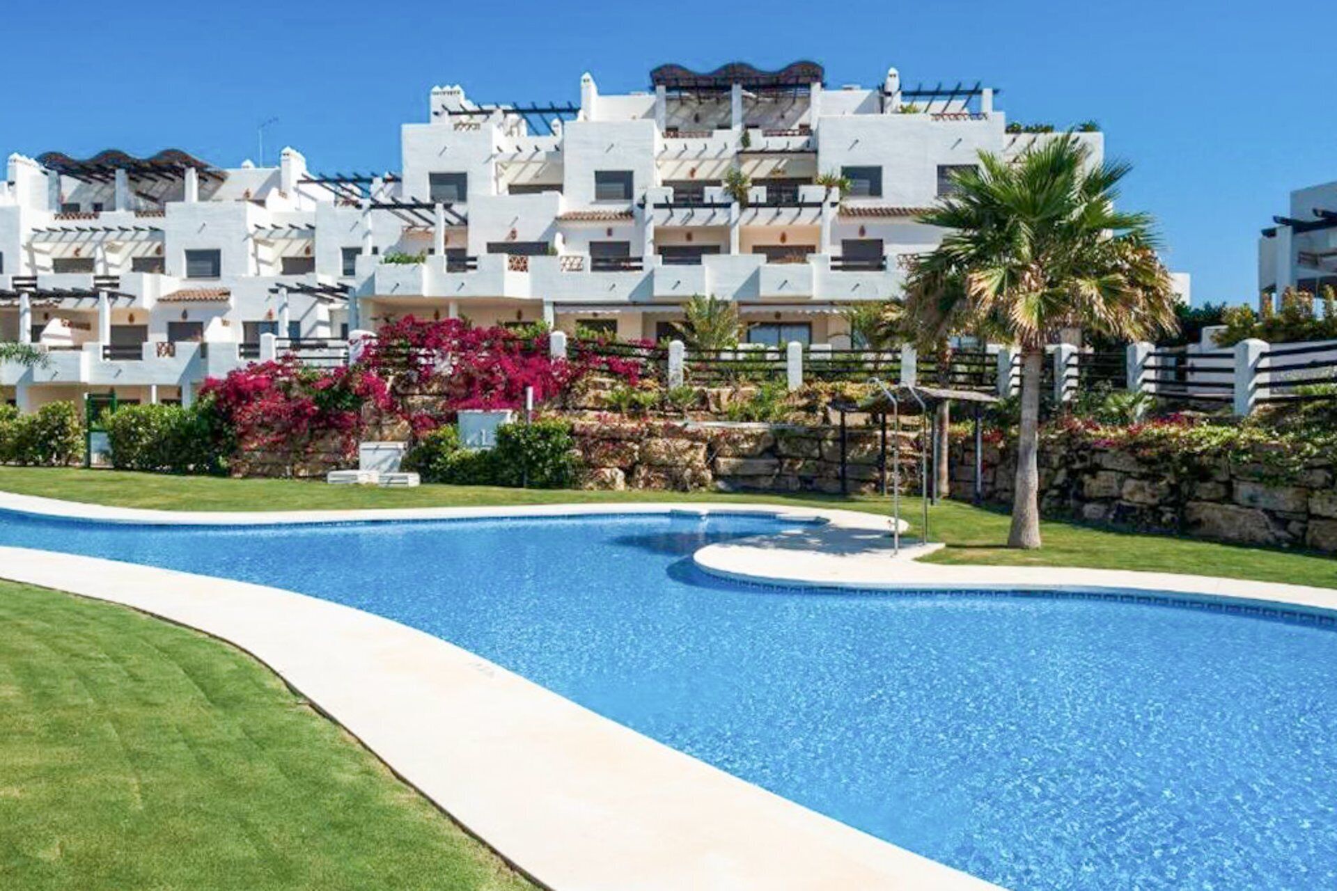 A swimming pool with a curved edge, surrounded by a well-maintained lawn and a row of white buildings with balconies.