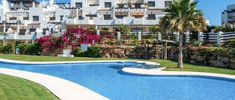 A swimming pool with a curved edge, surrounded by a well-maintained lawn and a row of white buildings with balconies.