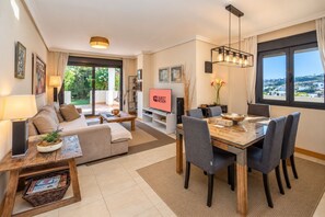 A modern living room with a sofa, coffee table, TV, dining table, and chairs. - GuestReady - Sunset Golf with Private Garden (Estepona)