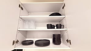 Ett vitt skåp med tre hyllor, där varje hylla innehåller svarta muggar, tallrikar och en genomskinlig plastpåse.
