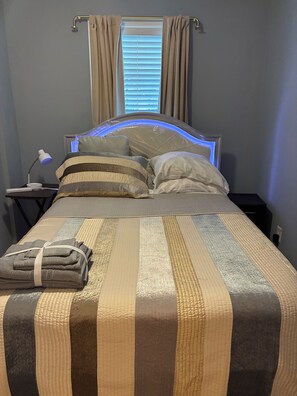 En snyggt bäddad säng med ett randigt överkast, en sänglampa och ett fönster med gardiner.