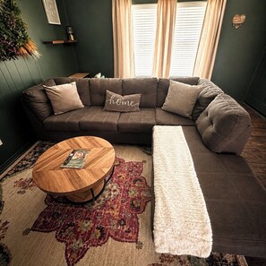 Een gezellige woonkamer met een hoekbank, een ronde houten salontafel en een kamerbreed tapijt met een patroon.
