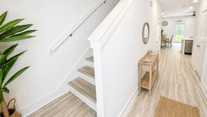 A staircase with white railings, a wooden table, and a plant in a hallway leading to a kitchen area. - Quick Drive to Beach | Sleeps 6 (Santa Rosa Beach)