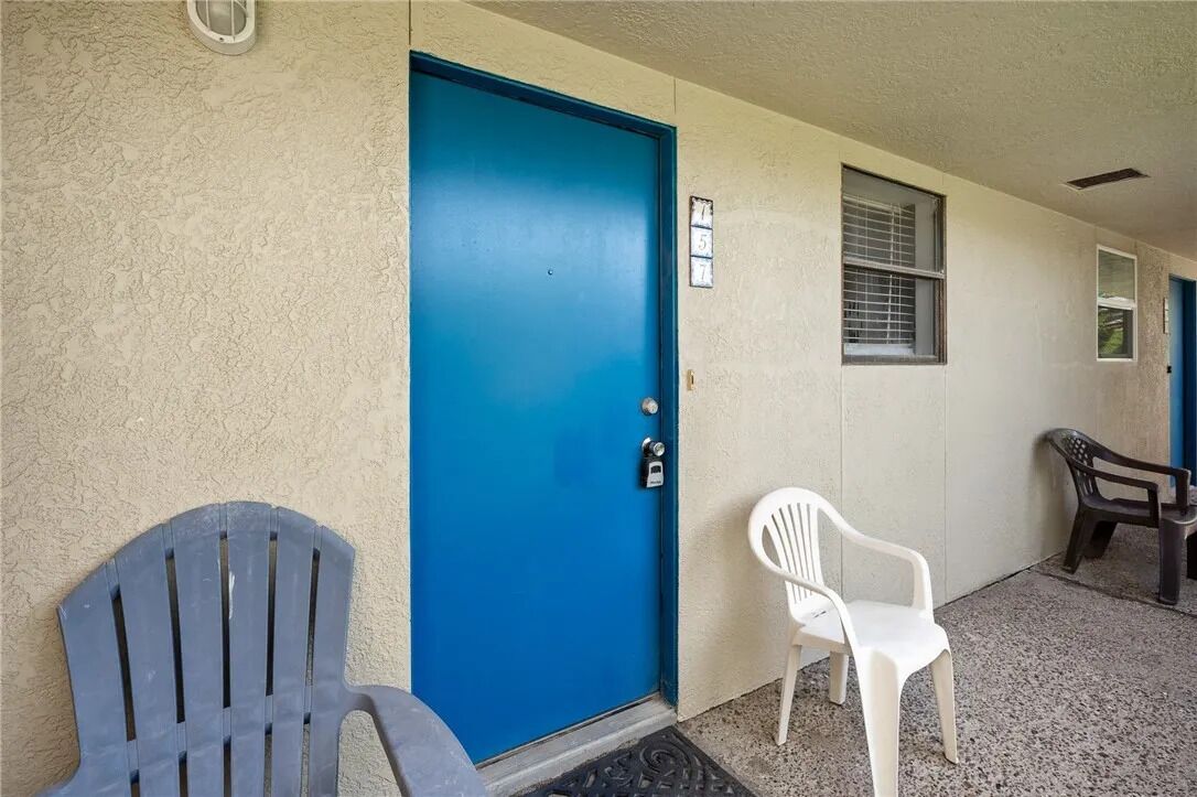 A blue door with a number sign, two plastic chairs, and a window with blinds.