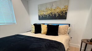 Um quarto com uma cama, um quadro, uma janela com persianas e uma mesa pequena.