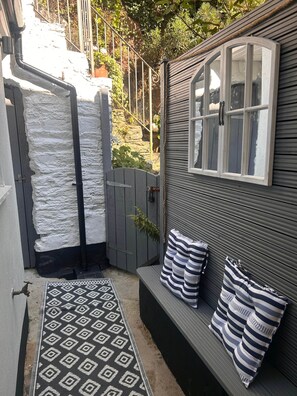 A small outdoor seating area with a bench and cushions, a gate, and a patterned rug. - The Snug at No 40 (Kingsbridge)