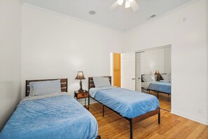 A bedroom with two beds, wooden flooring, a ceiling fan, and a door leading to another room. - Beachfront Oasis | 3BR | Ocean Views | Steps 2 HB (Huntington Beach)