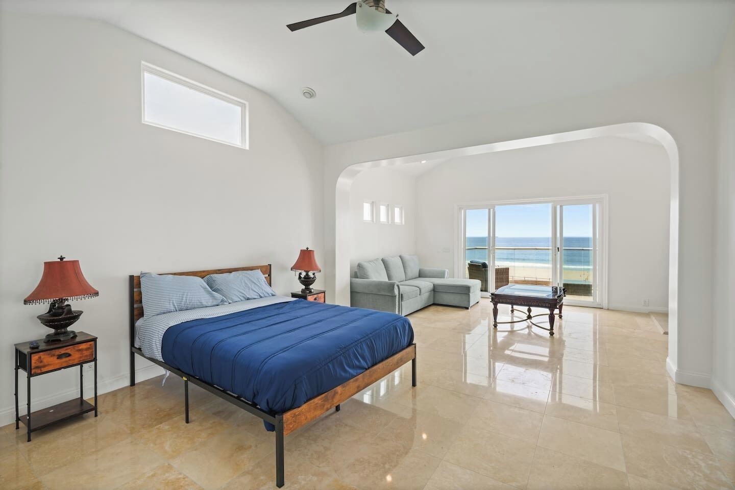 A bedroom with a bed, two bedside tables with lamps, a grey sofa, a small table, and a view of the beach through a glass door.