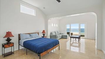 A bedroom with a bed, two bedside tables with lamps, a grey sofa, a small table, and a view of the beach through a glass door.