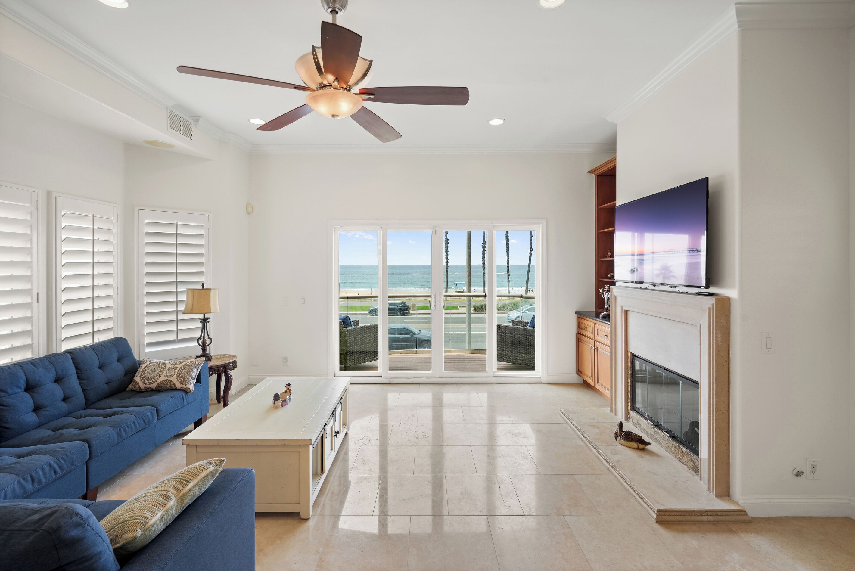 Uma sala de estar ampla com ventilador de teto, um sofá azul, uma mesa de centro, uma lareira e uma grande janela com vista para a praia.