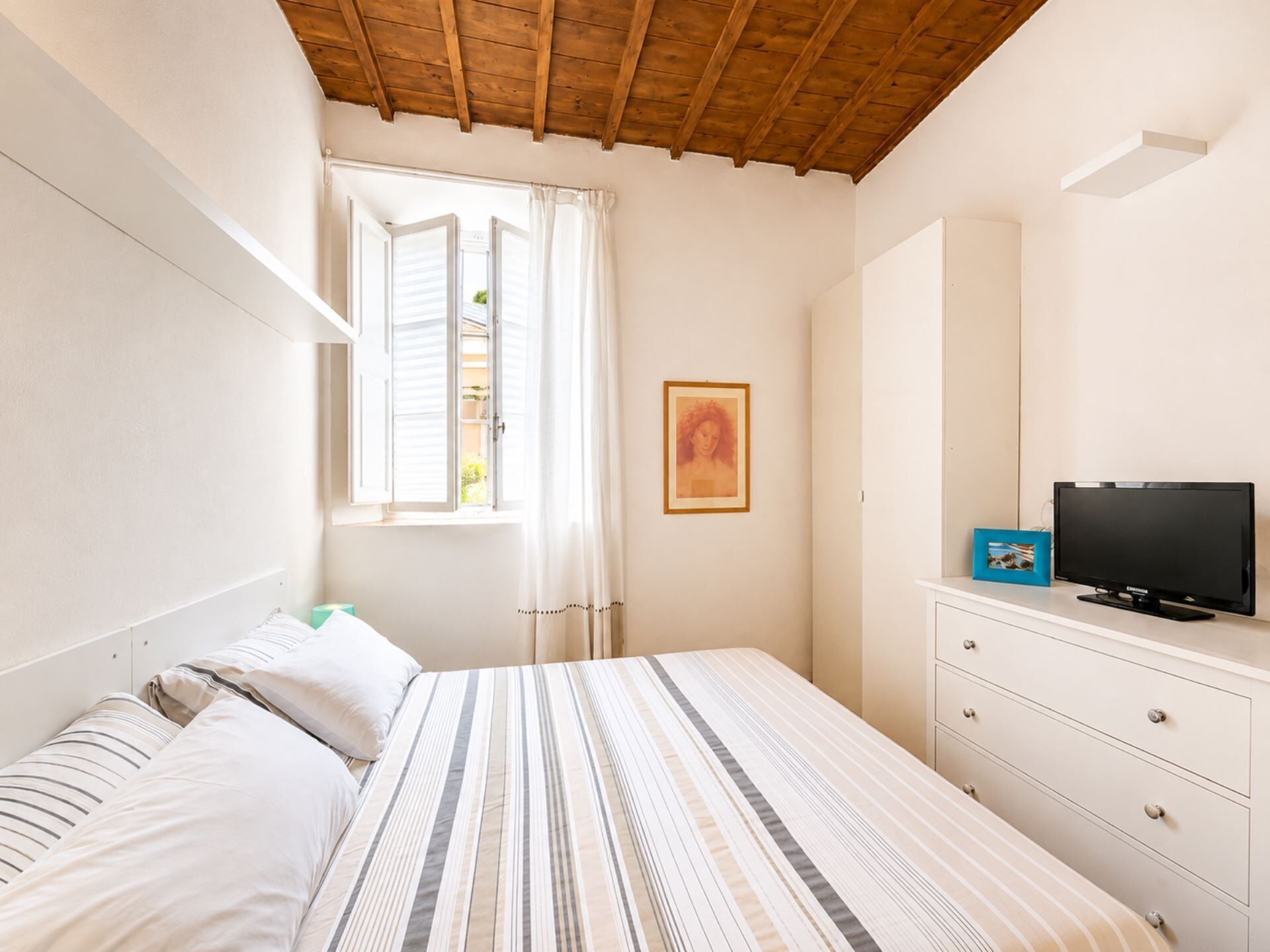 A bedroom with a bed, a TV, a dresser, a window with curtains, and a framed picture on the wall.
