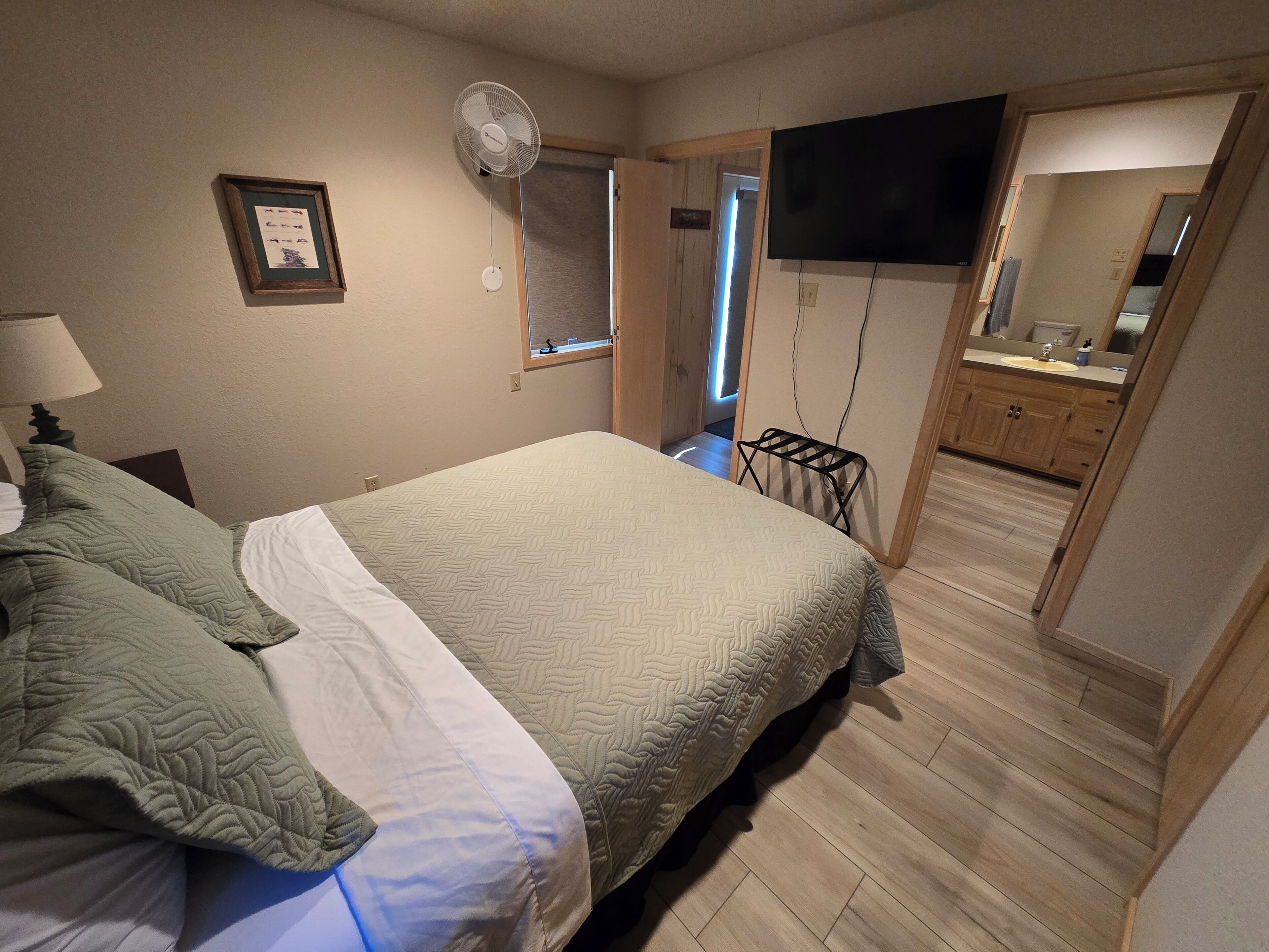 Un dormitorio con cama, mesita de noche, televisor montado en la pared, ventana y puerta que da a un baño.