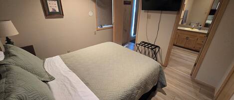 Un dormitorio con cama, mesita de noche, televisor montado en la pared, ventana y puerta que da a un baño.