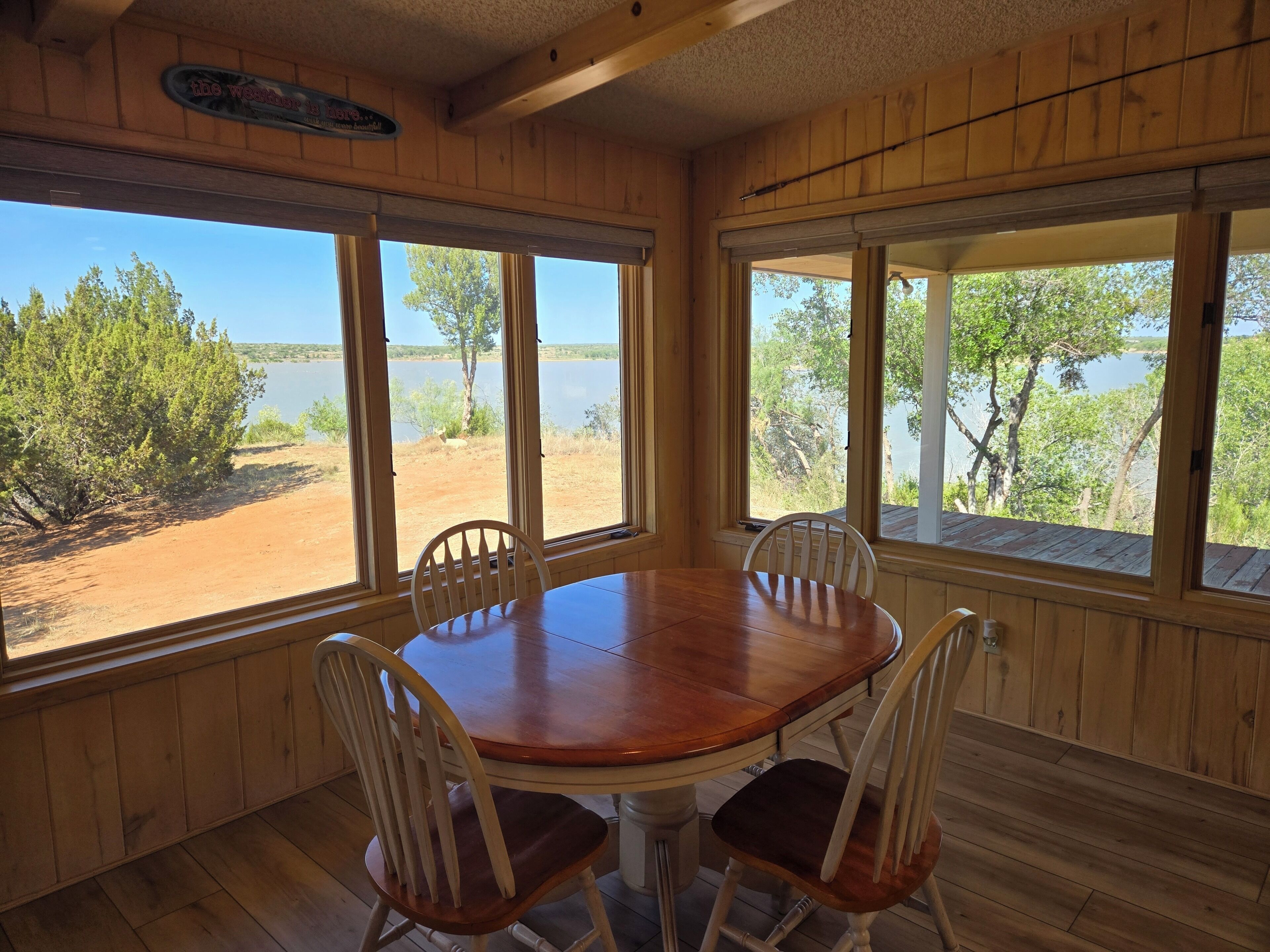 A wooden interior with a round table and four chairs, large windows with a view of a lake and trees, and a wooden ceiling.