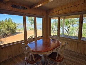 A wooden interior with a round table and four chairs, large windows with a view of a lake and trees, and a wooden ceiling.
