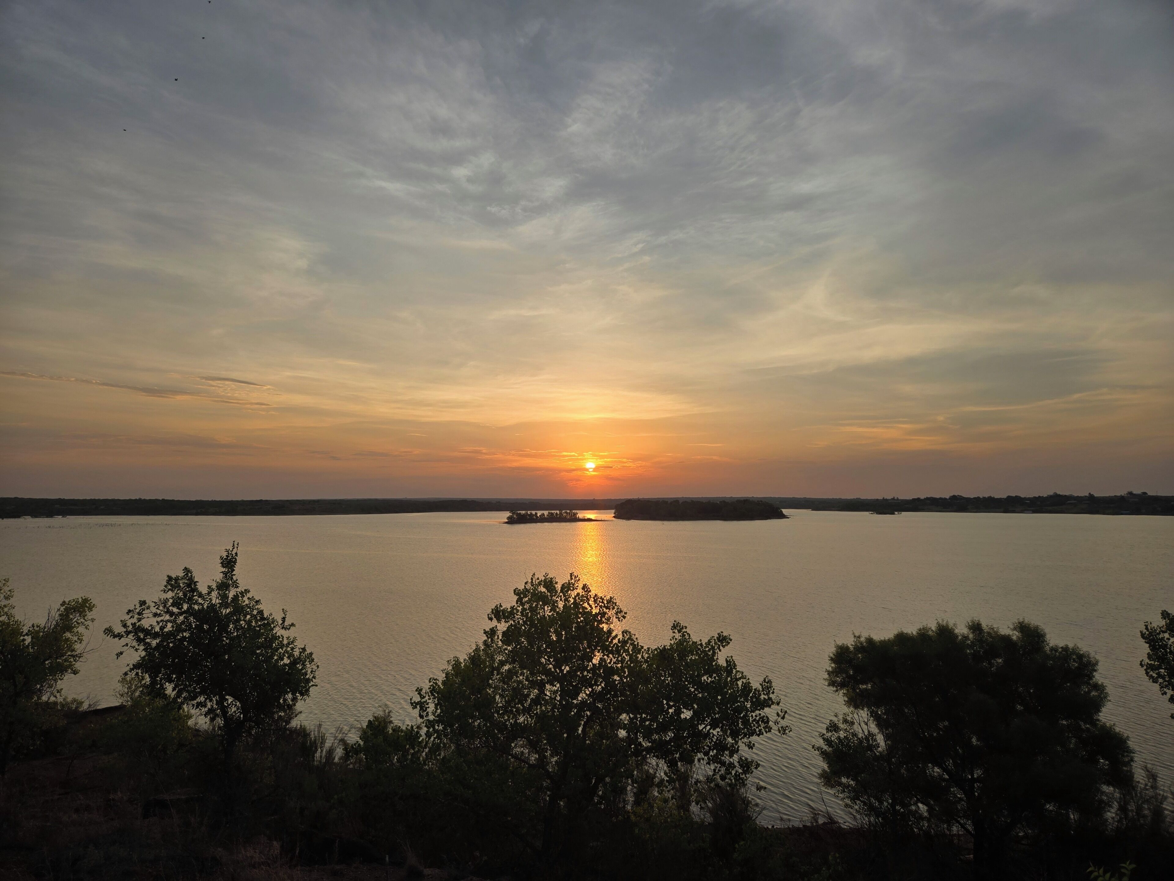 Ein Sonnenuntergang über ruhigem Wasser, im Vordergrund Bäume.