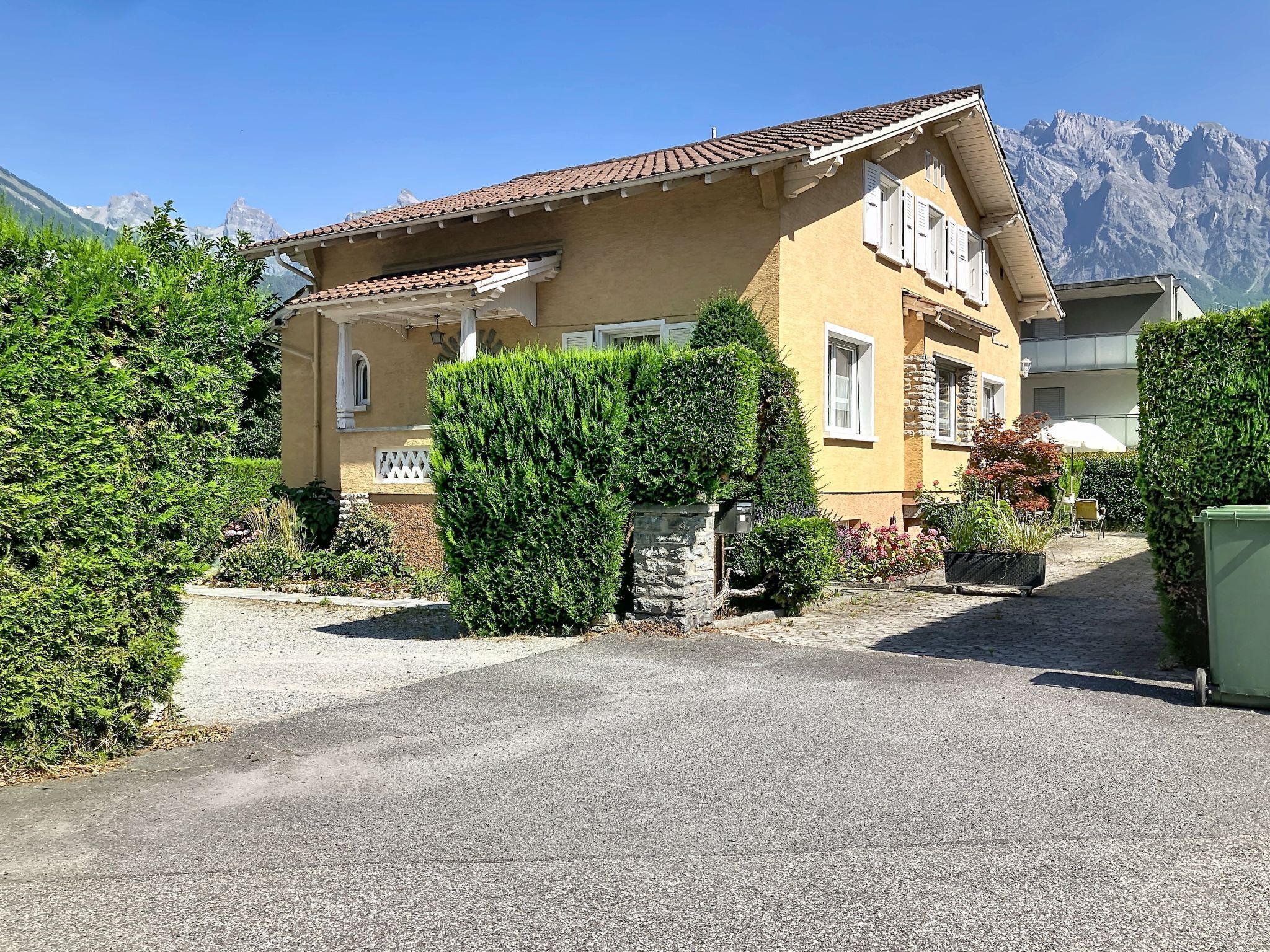 Un bâtiment jaune, surmonté d’un toit en tuiles, entouré de verdure et avec des montagnes en arrière-plan.