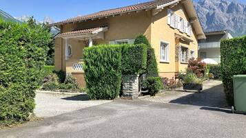 Un bâtiment jaune, surmonté d’un toit en tuiles, entouré de verdure et avec des montagnes en arrière-plan.