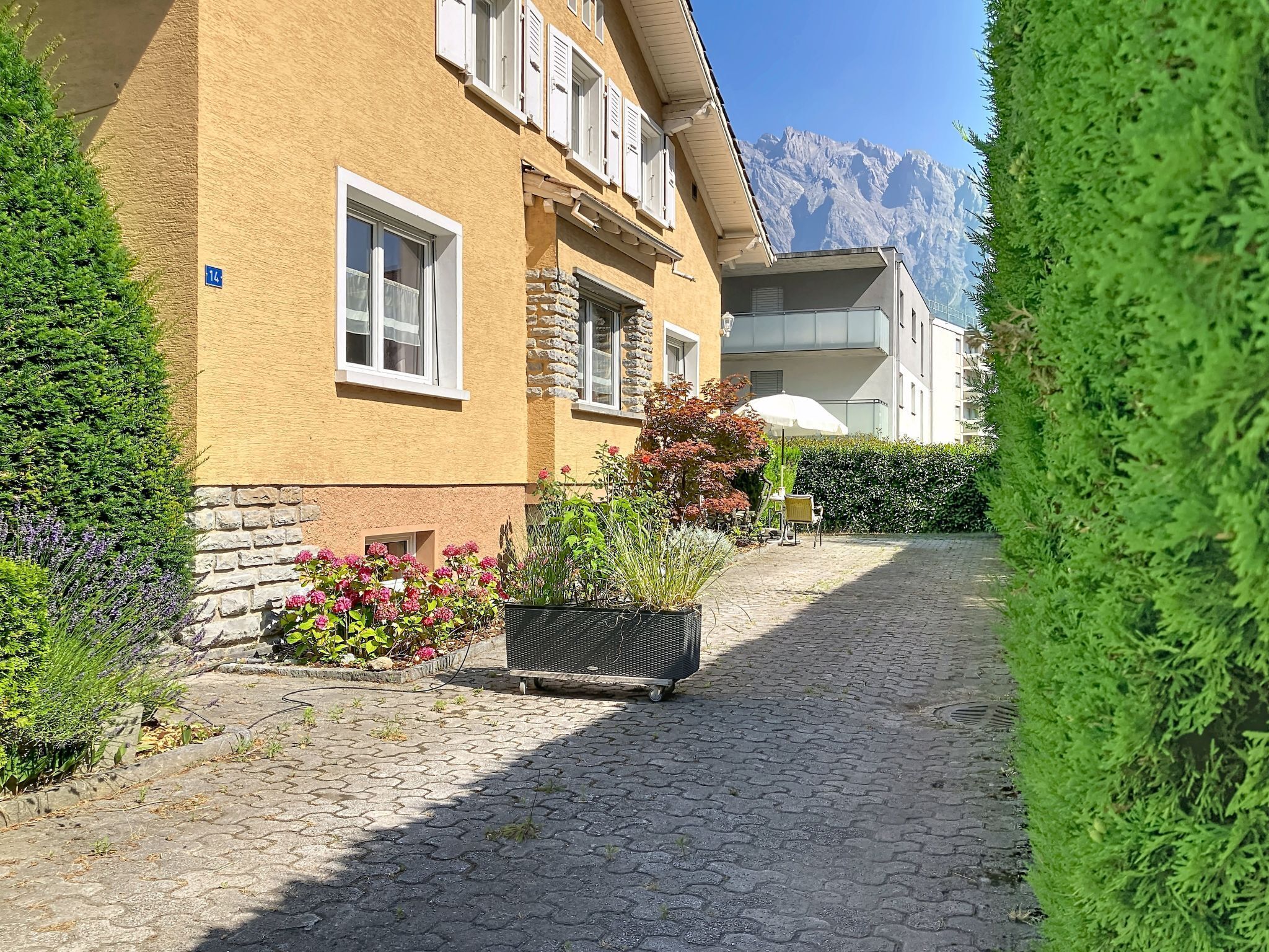 Un sentier pavé de pierres menant à un bâtiment jaune orné de détails en pierre, doté d’un balcon et offrant une vue sur la montagne.