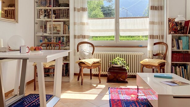 Una habitación con un ventanal grande, un radiador, una estantería, una mesa y dos sillas.