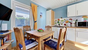 Una zona pranzo con tavolo e sedie in legno, una finestra con tende, una cucina con mobili bianchi e un televisore a schermo piatto su un supporto.