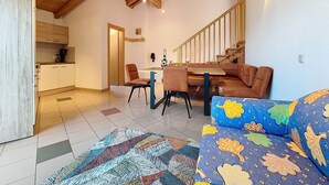 Un vaste espace de vie doté d’un plafond en bois, d’un canapé à motifs floraux, d’une table à manger avec des chaises, et d’une cuisine en arrière-plan.