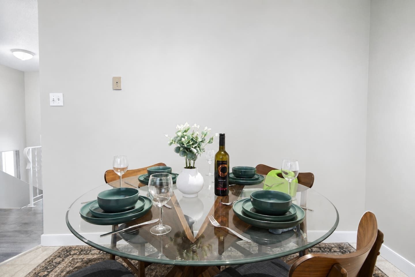 Un coin repas avec une table en verre, des assiettes vertes, un vase blanc rempli de fleurs et une bouteille de vin.