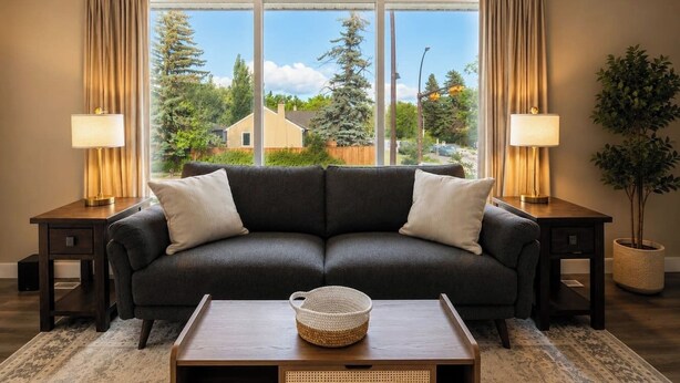 Ett vardagsrum med en mörk soffa, två sidobord med lampor, ett soffbord och utsikt över hus och träd genom stora fönster.