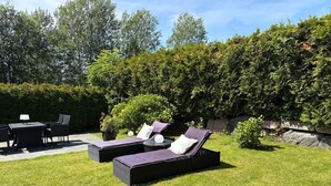 Een terras met tuinmeubilair, waaronder een tafel en stoelen, en twee loungestoelen met kussens.