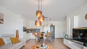 Una sala de estar moderna con un sofá, una mesa de centro y un televisor. Se ve una cocina al fondo con una zona de comedor.