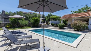 Piscina com espreguiçadeiras e um guarda-sol, cercada por vegetação e um edifício com telhado em azulejos.